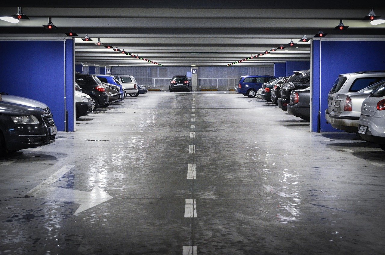 A well-lit underground parking lot with multiple rows of parked cars on both sides. The parking space is equipped with overhead lights, some of which have red indicators, likely showing occupied spaces. The floor is clean and reflective, with white arrows and lane markings guiding vehicles.