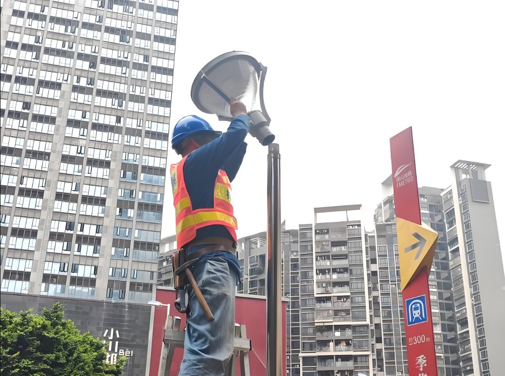 A technician wearing a safety helmet and reflective vest stands on a ladder while performing maintenance on an outdoor streetlight in an urban residential area, highlighting routine lighting upkeep tasks in city environments.