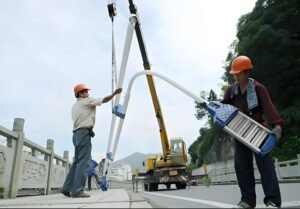 Two workers installing a large streetlight using a crane on a roadside, showcasing outdoor lighting maintenance and replacement